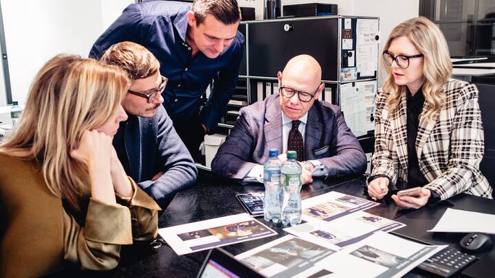Drei Männer und zwei Frauen sitzen an einem Tisch und besprechen anhand von Fotos den Stil der Chefs.