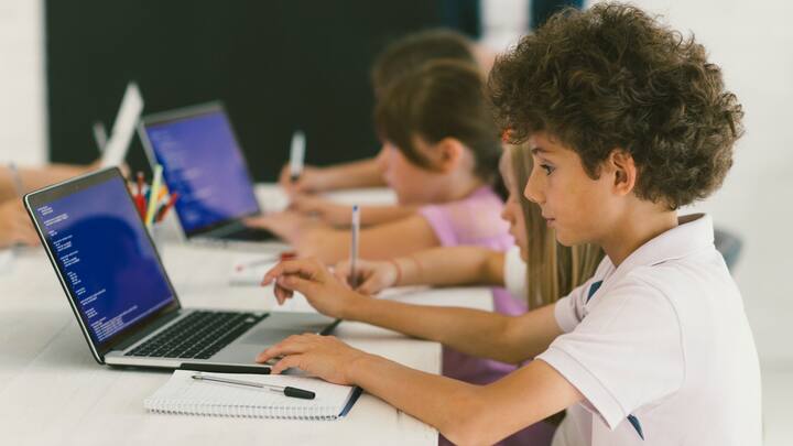 10 years old boy at coding class. Boy is sitting by the desk and using laptop, coding and taking notes. He is focus on his work. Kids in background sitting and coding.