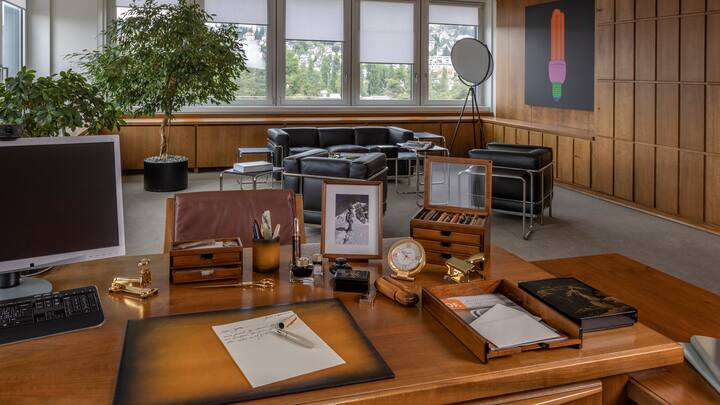 Schreibtisch mit diversen Schreibutensilien: Tintenfässer, Brieföffner, vergoldeter Locher, Schreibunterlage, Köcher aus Leder und ein Fotorahmen mit Bild eines Mannes in den Bergen.