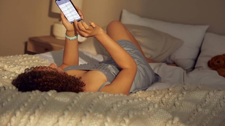 Frau liegt auf Bett mit Ava-Armbanduhr und schaut auf die Ava-App.