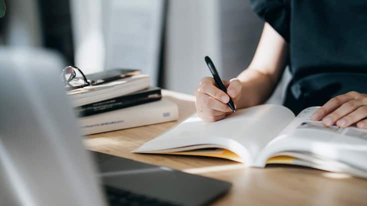 Cropped shot of focused young Asian woman reading book and making notes at home, concentrates on her studies. Further education, continuous learning concept
