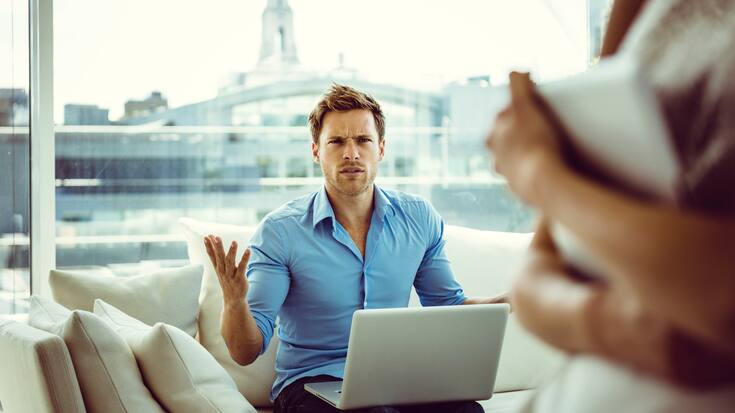Focus on young man sitting on sofa in the apartment with a laptop on his knees showing off irritation. 