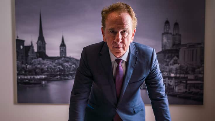 John Quinn im Zürcher Büro. Er steht vor einem Bild der Stadt Zürich und stützt seine Hände auf dem Tisch ab.