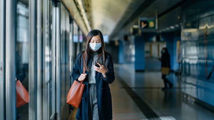 Asian woman with protective face mask using smartphone while walking on the subway platform