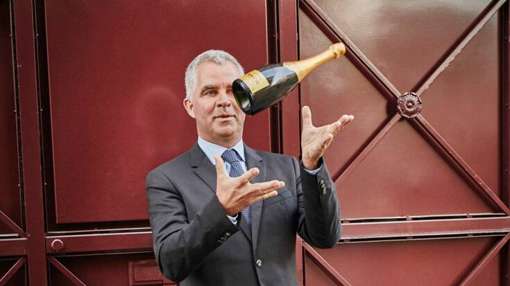 Champagner-Produzent Olivier Krug jongliert mit einer Champagnerflasche vor einem rustikalen Holztor.