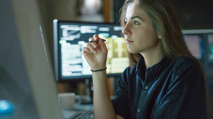 A young woman is seated at a desk surrounded by monitors displaying data, she is contemplating in this dark, moody office.