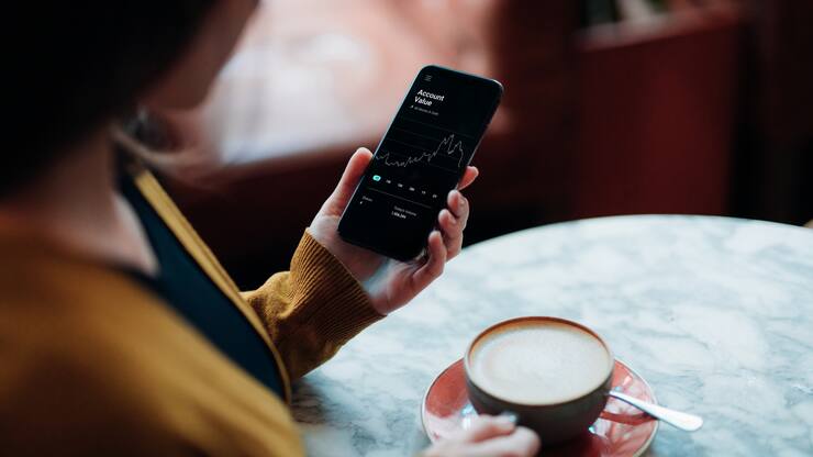 Junge Frau von hinten in Gelber Jacke in der linken Hand das Handy mit einem Kurschart und in der rechten eine Kaffeetasse.