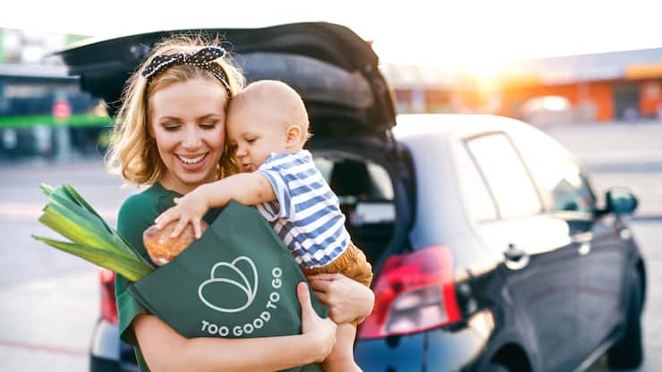 Beautiful young mother with her little baby son in front of a supermarket, holding paper shopping bag. Woman with a boy standing by the car.