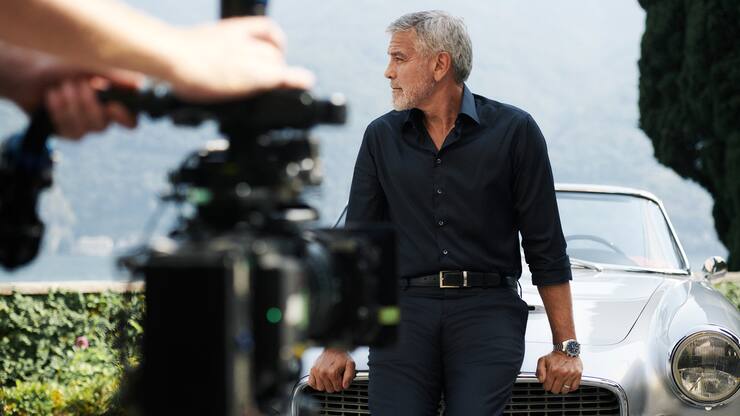 George Clooney ist einen silberfarbenen Maserati 3500 GT Spyder Vignale angelehnt und schaut auf die Seite.