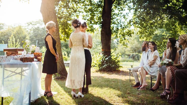 Lesbian couple holding hands in front of the altar. The wedding is outdoors and guest are seated in the foreground.