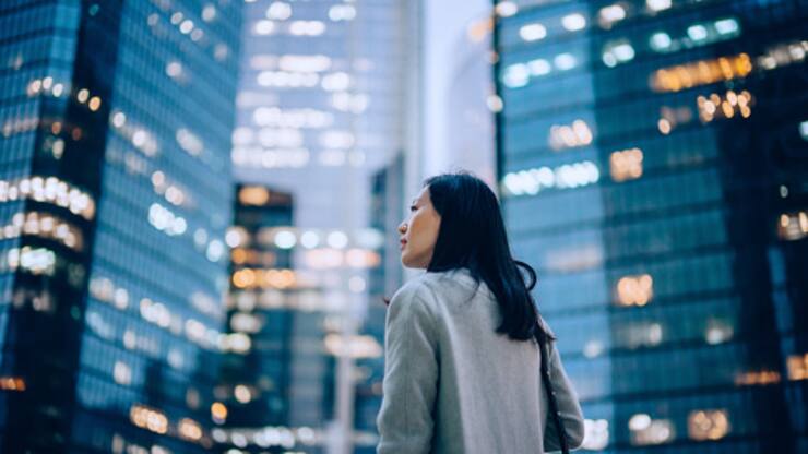 Low angle side profile of confident and professional young Asian businesswoman looking up while standing against contemporary corporate skyscrapers with illuminated facade in financial district in the evening. Female leadership and determined to success