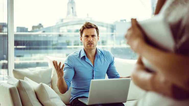 Focus on young man sitting on sofa in the apartment with a laptop on his knees showing off irritation. 