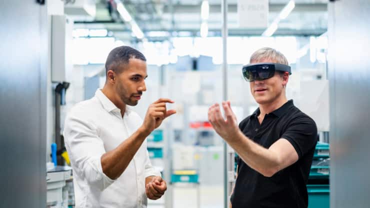 Technician wearing augmented reality glasses in a factory interacting with colleague 