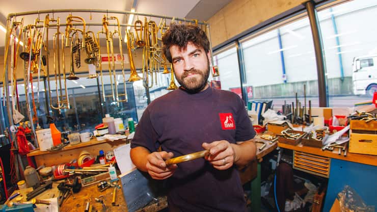 Fabian Bächi in seiner Werkstatt, er hält ein Baustück in der Hand, hinter ihm hängen Trompeten.