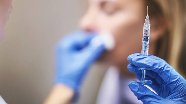 Unrecognizable woman having her lips and nasolabial folds done at private medical clinic. Syringe at focus.