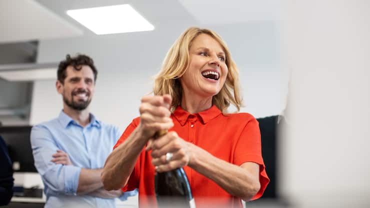 Cheerful mature businesswoman opening champagne bottle while celebrating success in creative office