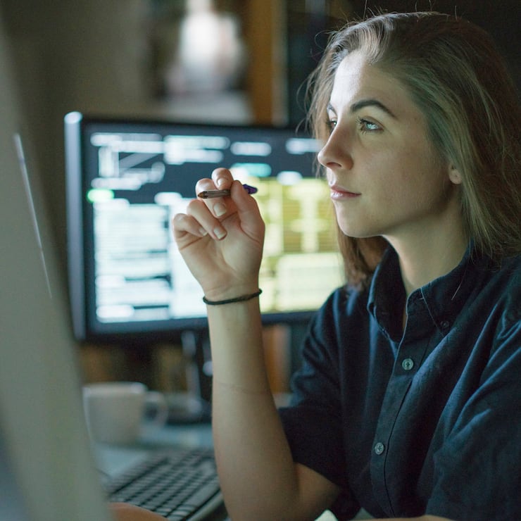 A young woman is seated at a desk surrounded by monitors displaying data, she is contemplating in this dark, moody office.