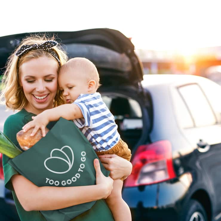 Beautiful young mother with her little baby son in front of a supermarket, holding paper shopping bag. Woman with a boy standing by the car.