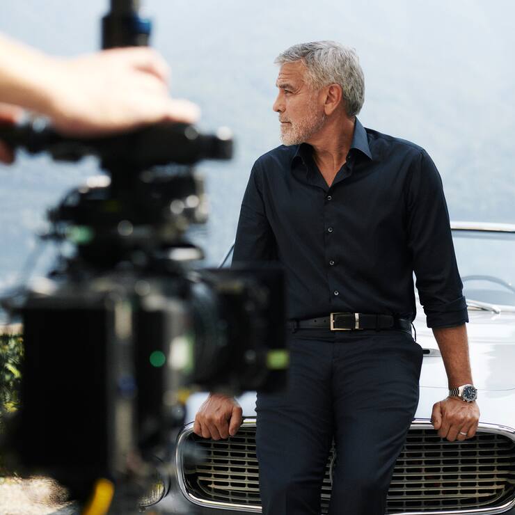 George Clooney ist einen silberfarbenen Maserati 3500 GT Spyder Vignale angelehnt und schaut auf die Seite.