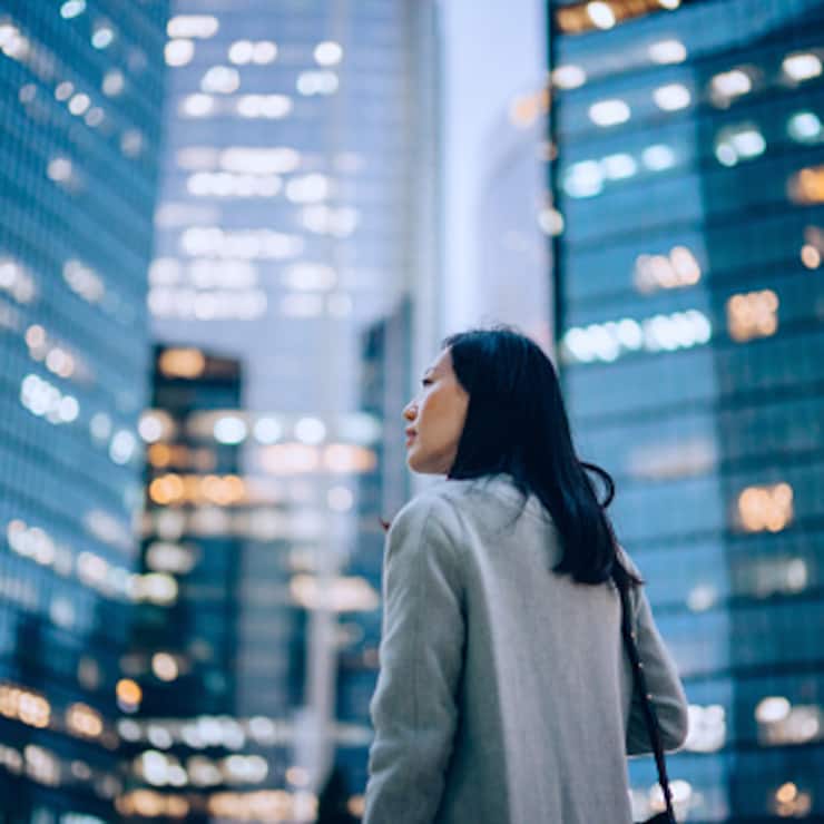 Low angle side profile of confident and professional young Asian businesswoman looking up while standing against contemporary corporate skyscrapers with illuminated facade in financial district in the evening. Female leadership and determined to success
