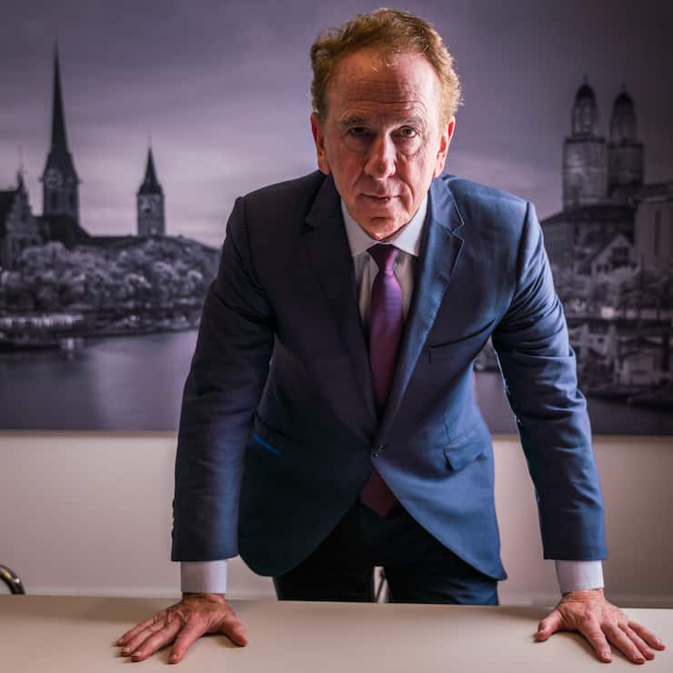 John Quinn im Zürcher Büro. Er steht vor einem Bild der Stadt Zürich und stützt seine Hände auf dem Tisch ab.