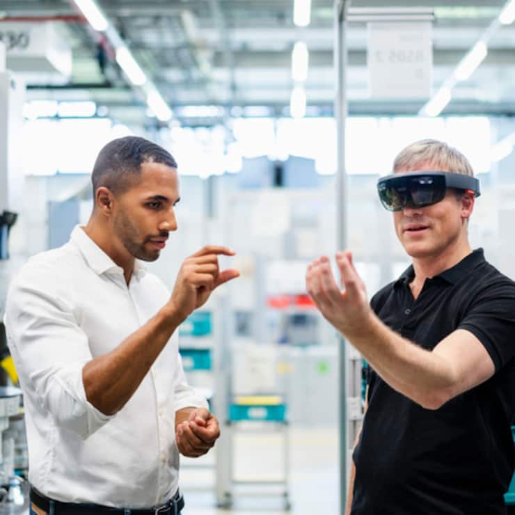 Technician wearing augmented reality glasses in a factory interacting with colleague 