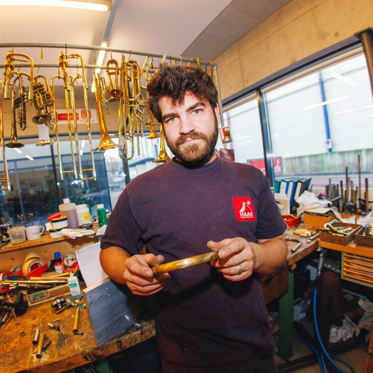 Fabian Bächi in seiner Werkstatt, er hält ein Baustück in der Hand, hinter ihm hängen Trompeten.