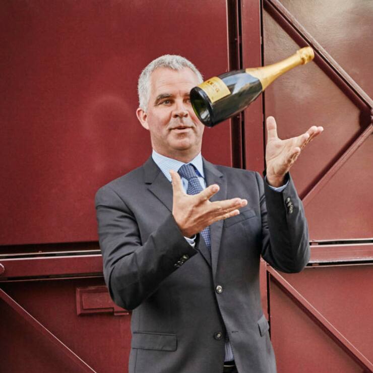 Champagner-Produzent Olivier Krug jongliert mit einer Champagnerflasche vor einem rustikalen Holztor.