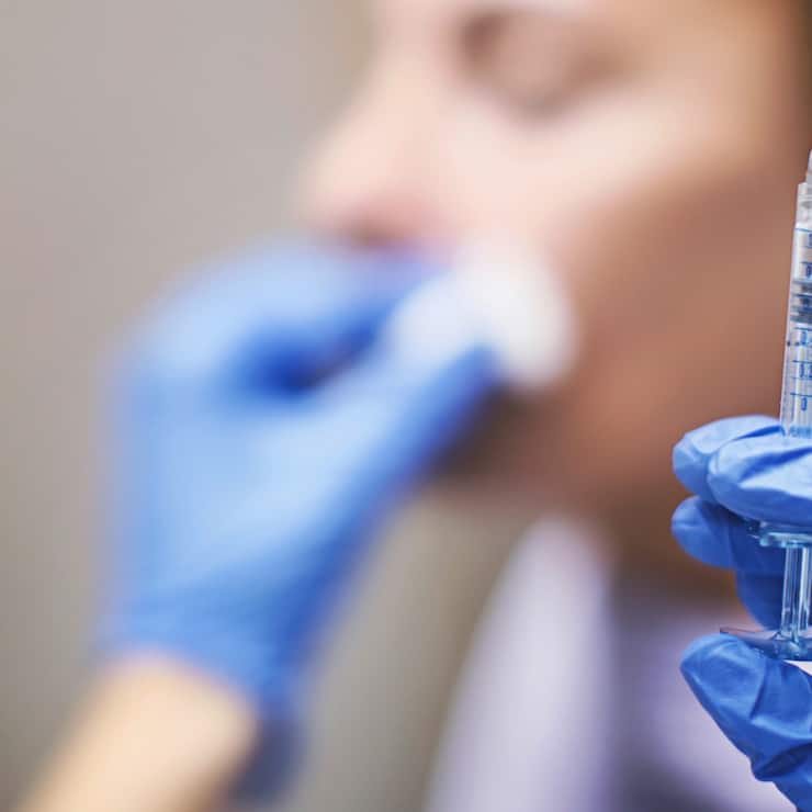 Unrecognizable woman having her lips and nasolabial folds done at private medical clinic. Syringe at focus.