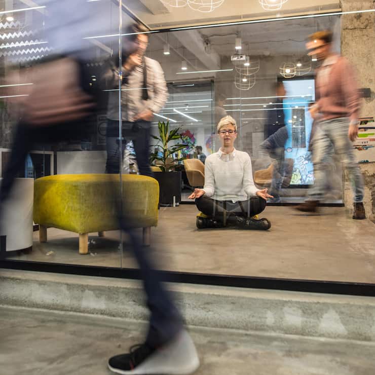 Young female entrepreneur meditating with eyes closed in busy office.