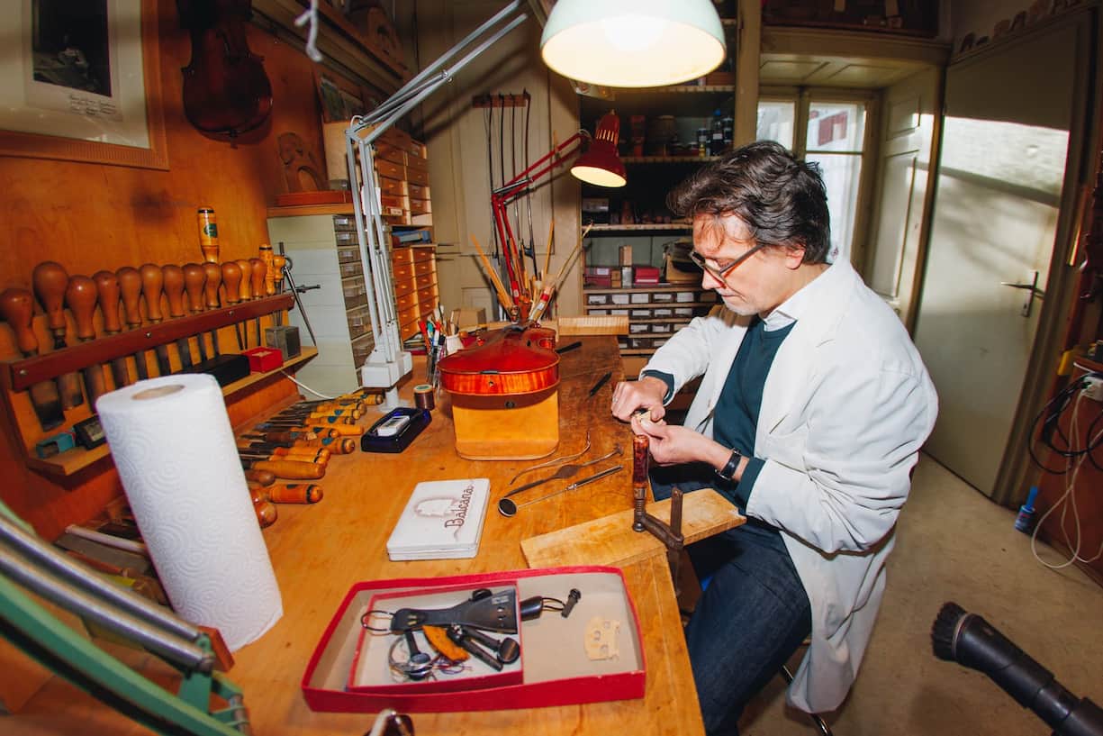 Der Geigenbauer sitzt am Werktisch und schnitzt Holz.