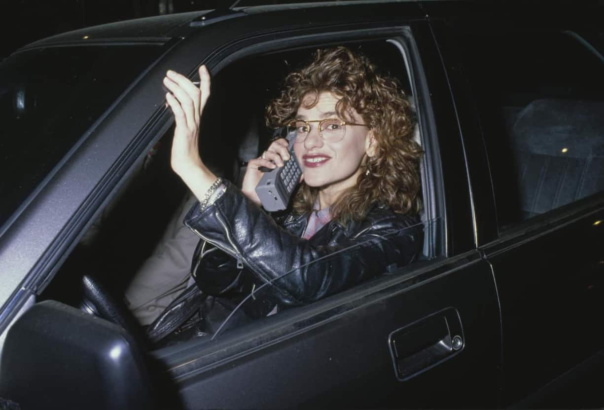 Mobiltelefonie, 1980Das Telefon gab es schon lange, auch das Mobilfunknetz gab es bereits seit 1958. Aber ab 1978 kam mit dem zellulären Mobilfunknetzneuer Schwung in die Mobiltelefonie. Bis aber jede und jeder wie Schauspielerin Sandra Bernhard ortsunabhängig und selbst am Lenkrad telefonieren konnte, sollten noch zwanzig Jahre vergehen. Handys brachten ­Veränderungen in der Arbeitsorganisation und im Management mit sich. Die Debatte um die «Work-Life-Balance» begann.