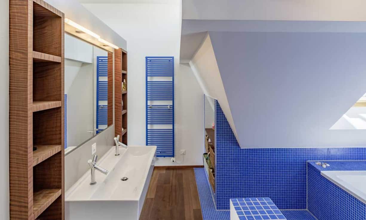 Das Badezimmer verfügt über zwei Waschtische sowie eine Badewanne und ist mit blauen Mosaiksteinen verkleidet. Gegenüber dem verglasten Bereich befinden sich ein farbenfrohes Gästebad mit Waschturm.