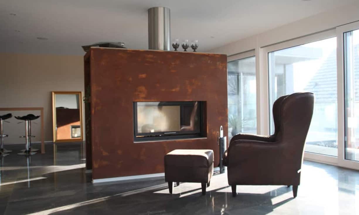 Das Warmluft-Cheminée trennt den Wohnraum architektonisch ab – und die doppelseitige Glasfront ermöglicht es, das Feuer von der Küche wie auch vom Wohnzimmer aus zu sehen.