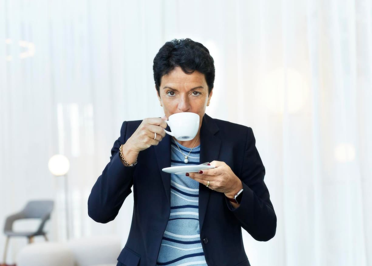 Porträt von Sabrina Soussan in schwarzem Blazer beim Kaffeetrinken.
