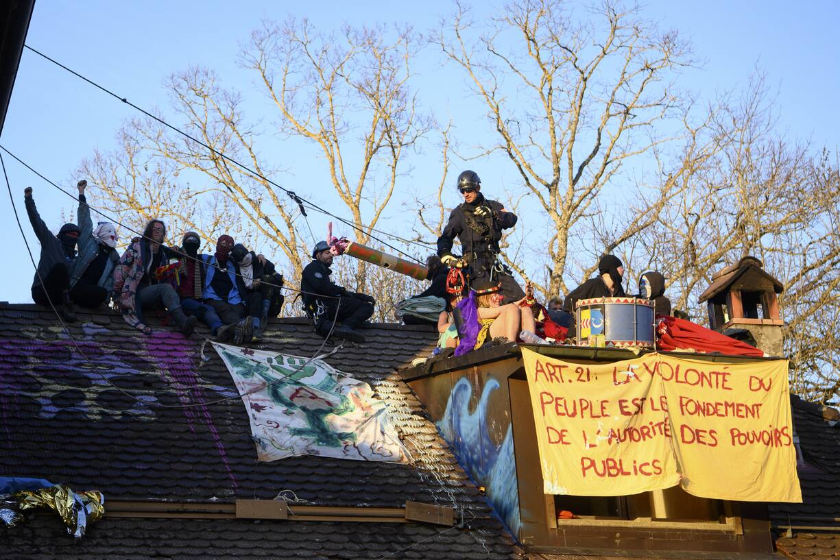 Environmental activists stand oin the roof of the house as Swiss police officers try to arrest them during the operation of the eviction of environmental protesters from the ZAD de la Colline "Zone A Defendre" (zone to defend) installed by environmental activists in the the so-called "plateau de la Birette" to block the extension of the Mormont quarry operated by the cement company LafargeHolcim between Eclepens and La Sarraz, Switzerland, Tuesday, March 30, 2021 (KEYSTONE/Jean-Christophe Bott)
