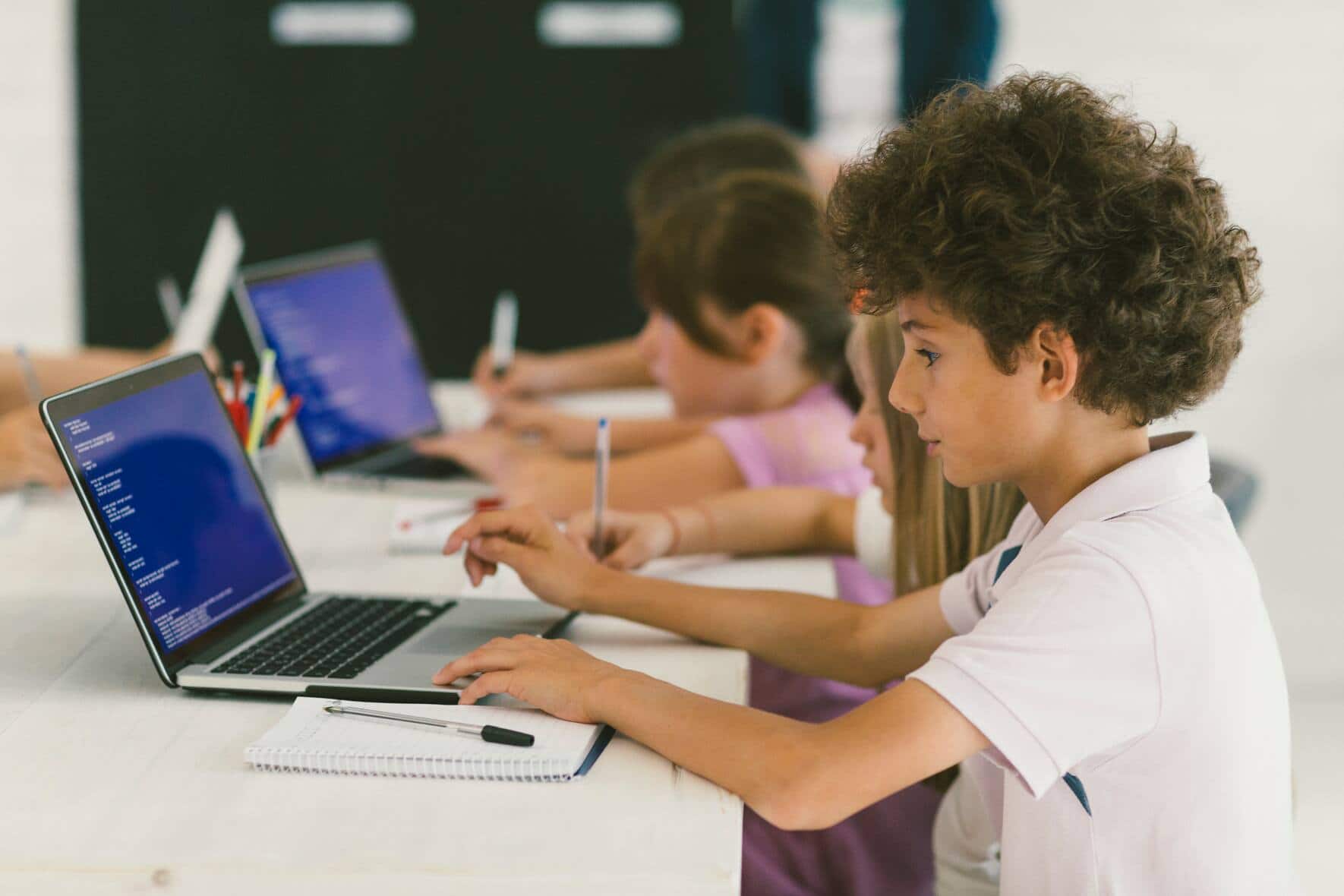 10 years old boy at coding class. Boy is sitting by the desk and using laptop, coding and taking notes. He is focus on his work. Kids in background sitting and coding.