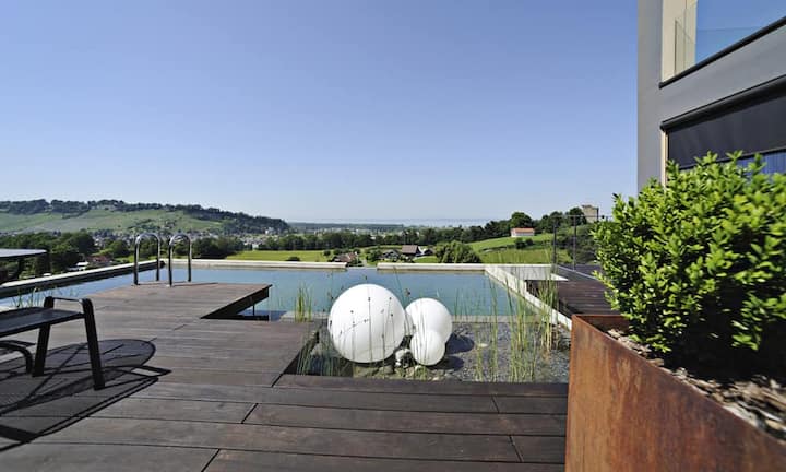 Die Terrasse besticht mit ihrem natürlichen, dunklen Holzboden, dem in den Pool integrierten Biotop mit Infinity-Kante. Von hier sowie auch...