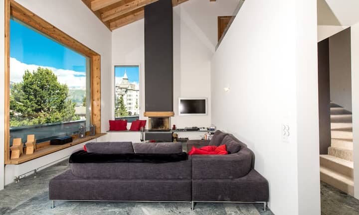 Der Wohnbereich mit Kamin besticht durch die besondere Raumhöhe und ermöglicht einen uneingeschränkten Blick auf das imposante Engadiner Bergpanorama.