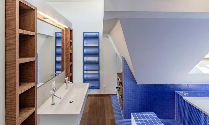 Das Badezimmer verfügt über zwei Waschtische sowie eine Badewanne und ist mit blauen Mosaiksteinen verkleidet. Gegenüber dem verglasten Bereich befinden sich ein farbenfrohes Gästebad mit Waschturm.