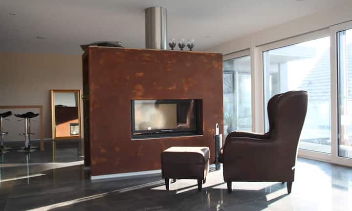 Das Warmluft-Cheminée trennt den Wohnraum architektonisch ab – und die doppelseitige Glasfront ermöglicht es, das Feuer von der Küche wie auch vom Wohnzimmer aus zu sehen.